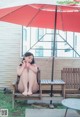 A naked woman sitting on a bench under an umbrella.